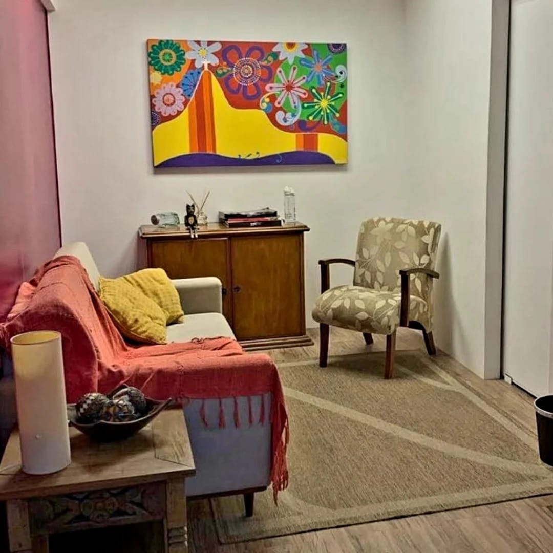 Consultório de Carla Espinola, com poltronas, tapete e uma pintura colorida de flores na parede
